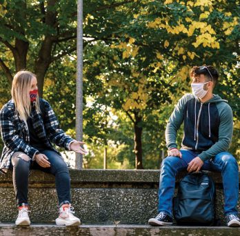A girl and boy student sit outside and talk in the fall with masks on.
                  