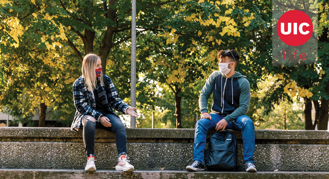 A girl and boy student sit outside and talk in the fall with masks on.