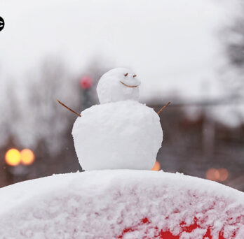 A snowman perched on a red half circle with tree line and traffic lights in the background
                  