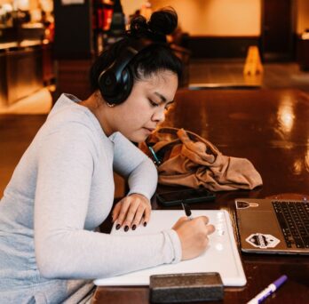 Student sitting at a desk writing on a notepad with headphones on.
                  