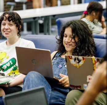 Students smiling while working on their laptops in the Commuter Student Resource Center.
                  